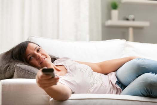 long haired smiling woman on a choush in her living room changing channels with a remote control