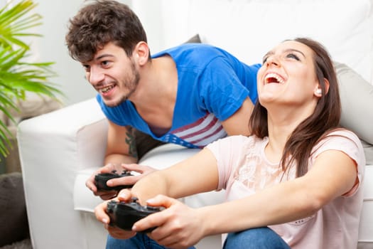 Laughing young couple playing video games on the sofa at home in a close up view with focus to the man as he leans forwards focused on the screen
