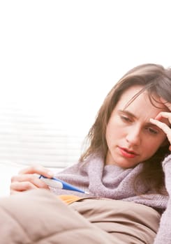 headache and illness of a young woman on a couch in her living room checking a termometer