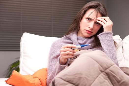 headache and illness of a young woman on a couch in her living room checking his termometer
