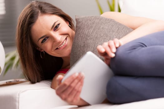 portrait of a smiling woman (in focus) with a digital tablet (blurred) lying on a sofa in her living room