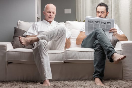 Father and son reading business news from a printed newspaper while sitting on a comfortable couch at home