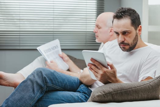 Man sitting alongside his elderly father on the sofa reading on a tablet with a look of absorbed concentration