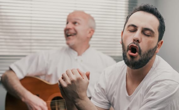 Man singing while his senior father or friend is playing the guitar as a hobby or a funny relaxing activity at home
