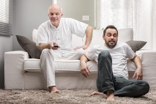 Two men sitting watching a TV program, one a senior man with an interested expression, the other younger showing bored indifference
