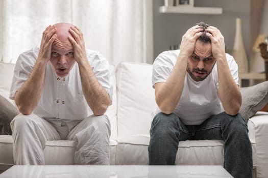 Senior father and adult son holding their head terrified by bad news or negative thinking sitting together on the couch at home