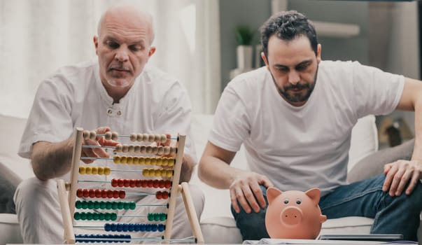 Senior man using an abacus to do financial calculations as his son sirs staring at his piggy bank