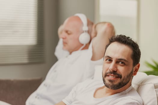 Man sitting smiling at the canera as his father or partner sits alongside him listens to music on headphones