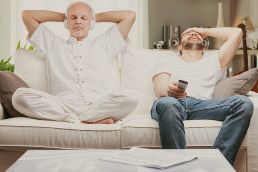 Two men relaxing at home, one older quietly meditating while listening to music and one younger laughing uproriously at a TV program