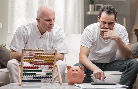 Two generations of men sitting doing financial calculations, the senior man using an abacus and the younger man a calculator