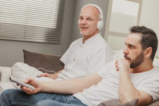 Senior man relaxing on a sofa with his son or partner listening to music on headphones and turning to smile quietly at the camera with a look of contentment