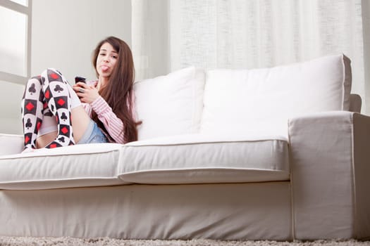 rascal girl showing her tongue stuck out as a joke while using her mobile phone in her living room on a couch