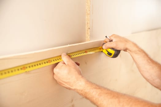 Unidentifiable man using long yellow tape measure on large wood plank leaning against white wall