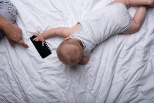 Little newborn baby reaching for a mobile phone touching the screen in a tactile gesture with the hand of his loving father close by on a white bed from above