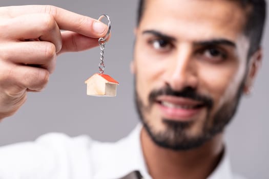 Smiling man holding up a key ring with miniature house in a concept of a dream home, ownership, purchase or sale or real estate with focus to his hand