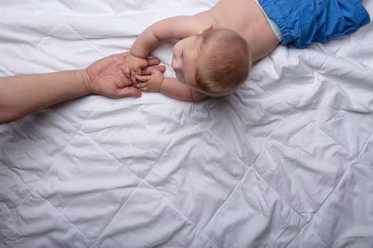 Moment of love and tenderness between a young father and his newborn baby son as they touch hands while lying together on a white bed viewed from above with copy space