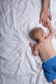 Little baby boy reaching out to touch its father on the hand as it lies on its stomach on a white duvet in a concept of love, bonding and closeness