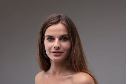 Young woman looking calmly at the camera with big brown eyes and a quiet smile as she poses with bare shoulders against a grey studio background