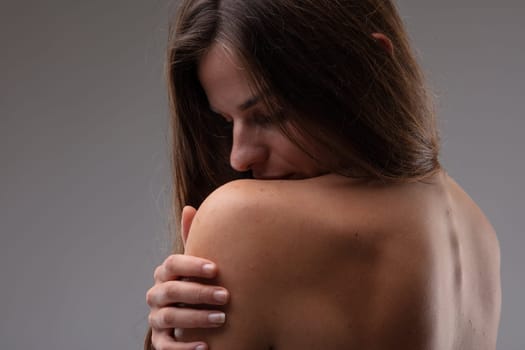 Seductive sensual woman caressing her bare shoulder with a hand as she turns back to the camera with closed eyes and a blissful expression on a grey studio background