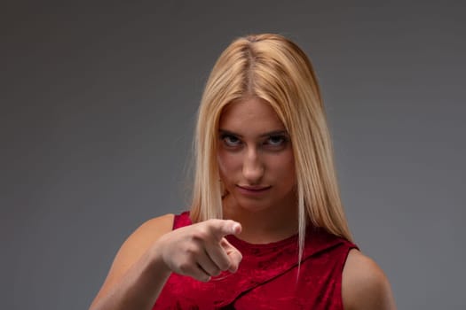 Stern glamorous blond woman pointing at the camera in a concept of blame, identification of choices on a grey studio background with copyspace