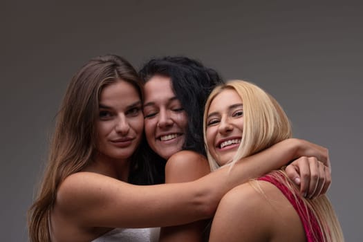 Three happy smiling attractive young woman arm in arm posing with their heads close together grinning at the camera against a grey studio background