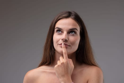 Young lady standing deep in thought with finger to lips looking up with a contemplative serious expression to blank copyspace in a head and shoulders portrait against a grey studio background