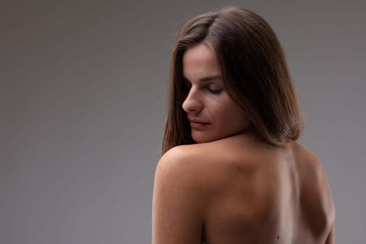 Seductive sensual topless young woman looking back over her bare shoulder and back with closed eyes and serene expression against a grey studio background
