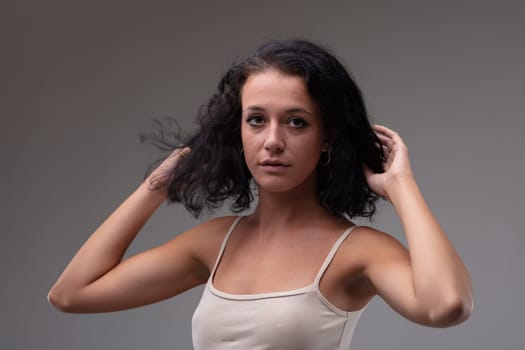 Cute young woman with curly shoulder length hair looking thoughtfully at the camera with a solemn expression and hands to head in a head and shoulders portrait against a grey studio background