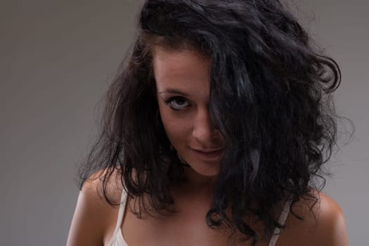 Seductive young woman with hair covering one eye and head bowed looking at the camera with a quiet smile in a close up cropped head and shoulders portrait against a grey studio background