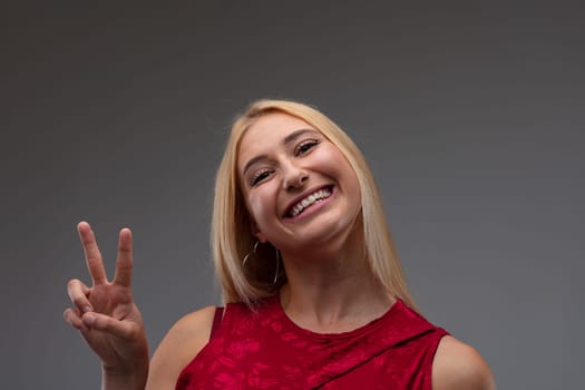 Cute charismatic blond woman in stylish red top giving a V-sign with a vivacious beaming smile of success on a grey studio background