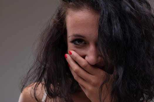 Young woman sniggering or giggling into her hand in a close up cropped portrait with her long hair falling over one eye on a grey studio background