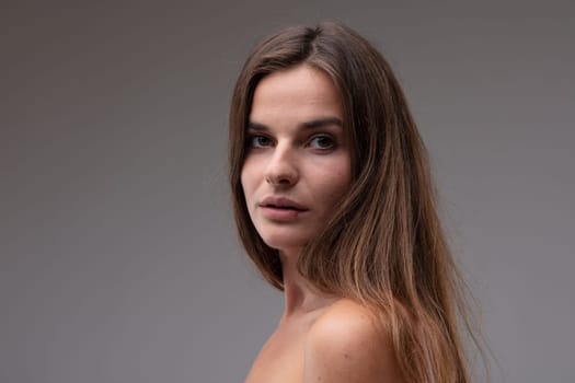 Natural portrait of a sincere young woman with naked shoulders looking back over her shoulder at the camera with wide eyes and parted lips in a head and shoulders portrait against a grey studio background