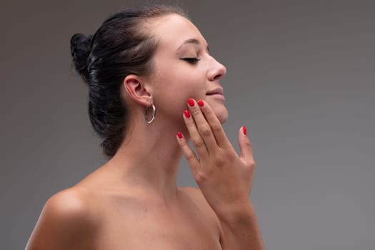 Sensual young woman with bare shoulders standing caressing her cheek with her hand with eyes closed in pleasure in a head and shoulders portrait against a grey studio background