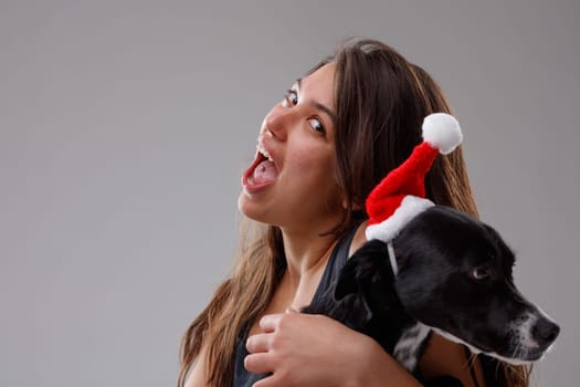 Young woman and her pet dog celebrating Christmas in festive red outfits as she hugs it in her arms while displaying the piercing in her tongue to the viewer