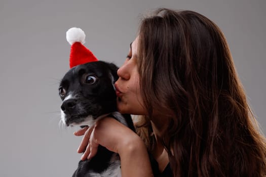 Loving young woman kissing her little pet dog wearing a festive red Santa hat for Christmas as she hugs it in her arms