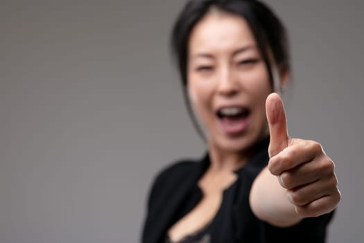 Excited happy Asian woman giving a thumbs up gesture of approval, excellence, success or agreement with focus to her hand over a grey studio background with copyspace