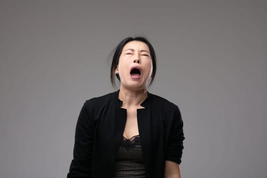 Woeful Asian woman wailing in anguish with her mouth open and a sorrowful expression over a grey studio background with copyspace
