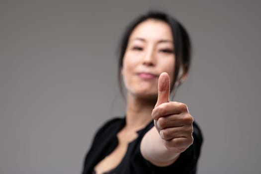 Elated triumphant Asian woman giving a thumbs up gesture of success and approval with focus to her hand over a grey studio background with copyspace