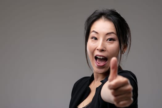 Enthusiastic Asian woman cheering giving a thumbs up gesture of success and achievement over a grey studio background with copyspace with focus to her face