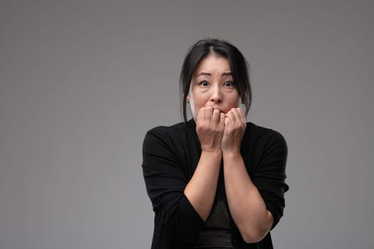Petrified Asian woman staring at the camera in panic biting her nails in her fear over a grey studio background with copyspace
