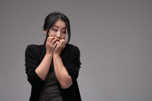Fearful anxious Asian woman looking to the side biting her nails in trepidation over a grey studio background with copyspace
