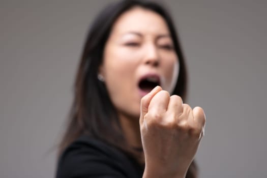 Aggressive Asian woman threatening with her fist and yelling with focus to her hand in close up over grey
