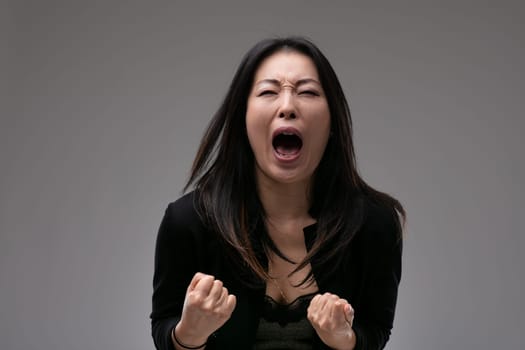Agitated upset woman yelling in anguish or frustration with clenched fists over a grey studio background with copyspace