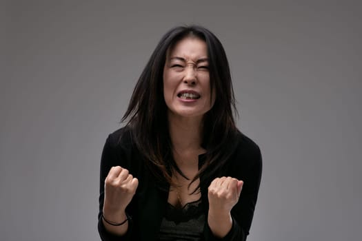 Upset emotional Asian woman clenching her fists in anguish as she implores the viewer or is overcome by loss against a grey studio background with copyspace