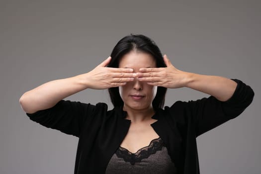 Asian woman in a smart black outfit covering her yes with her hands in a concept of the metaphor see no evil, hear no evil speak no evil, over a grey studio background