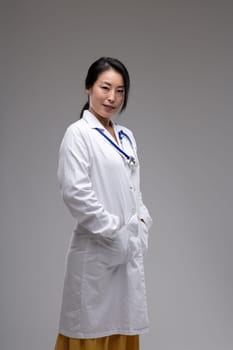 Thoughtful female Asian doctor or nurse in lab coat standing sideways looking t the camera with a quiet friendly smile over a grey studio background with copyspace