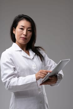 Female Asian doctor looking quizzically at the camera as she holds a clipboard in her hand pointing to the notes over grey