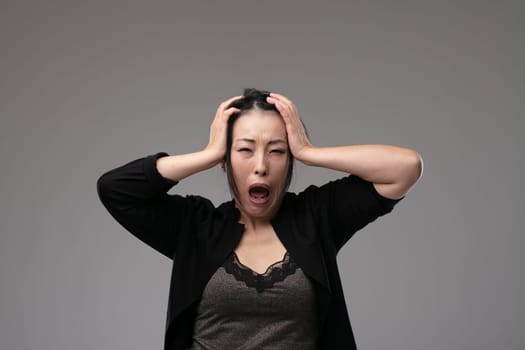 Sorrowful grieving Asian woman wailing in anguish holding her hands to her head over a grey studio background with copyspace