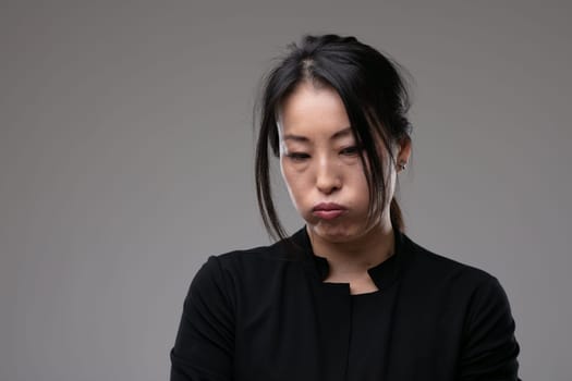 Petulant sulky Asian woman pouting her lips and looking down with a morose expression over a grey studio background with copyspace
