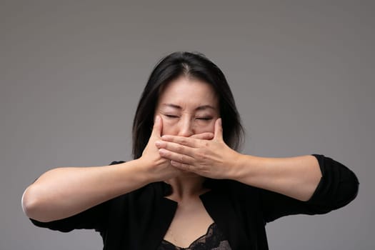 Asian woman with closed eyes covering her mouth with her hands in a concept of the metaphor see no evil, hear no evil speak no evil, over a grey studio background